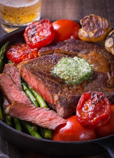 Pan fried ribeye steak in a cast iron skillet with roasted tomatoes, garlic head and asparagus.